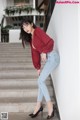 A woman in a red blouse and jeans leaning against a wall.